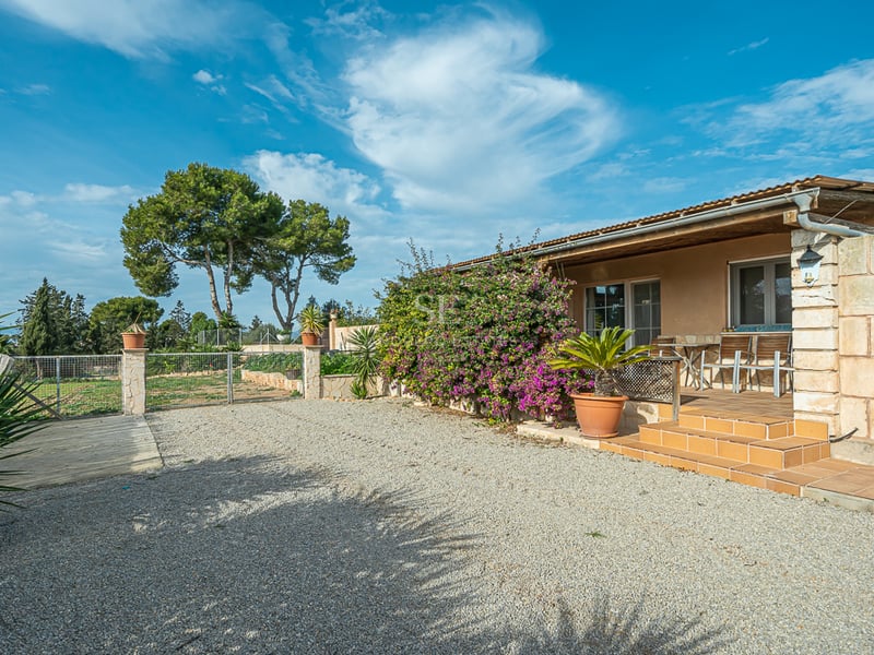 Façade en pierre d'une villa avec porche en terre cuite, cour en gravier et bougainvilliers sous un ciel bleu.