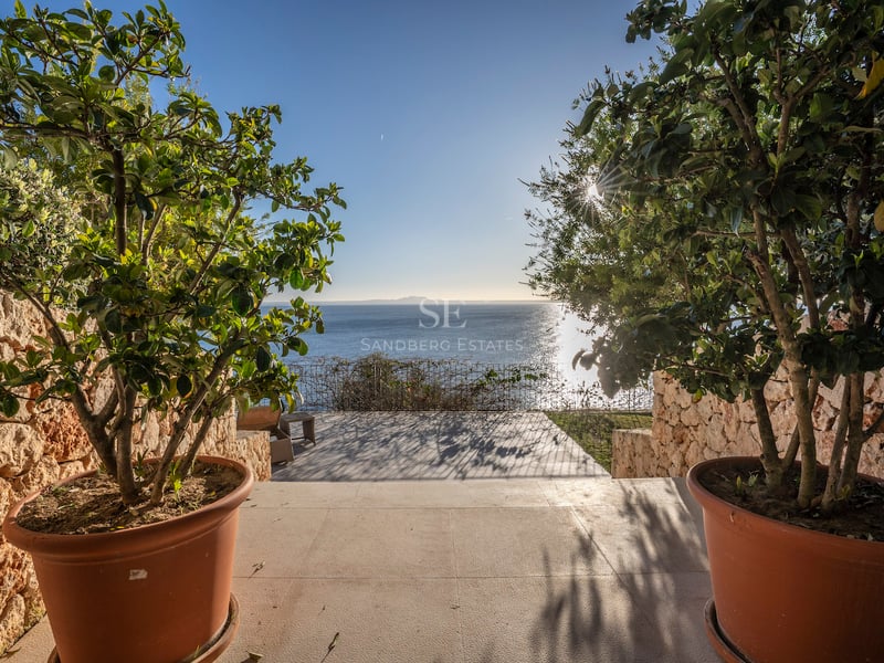 Blick auf das blaue Meer von einer Steinterrasse mit Topfpflanzen und Natursteinmauern unter blauem Himmel.
