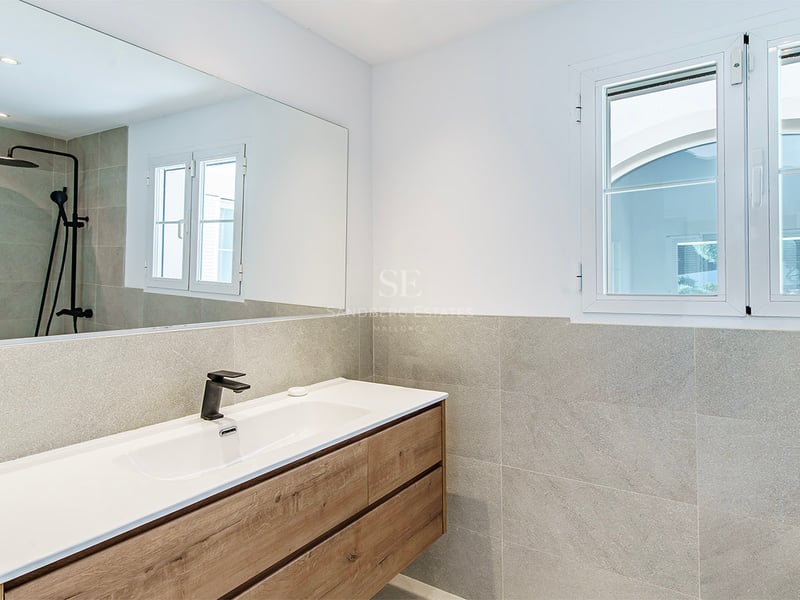 Modern badrumsinredning med träkommod, vitt tvättställ, svarta kranar och grått kakel.