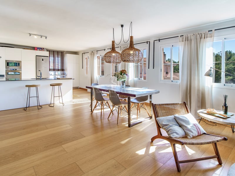 Soligt rum med öppen planlösning, vitt kök, matbord i trä och korglampor över ekparkett.