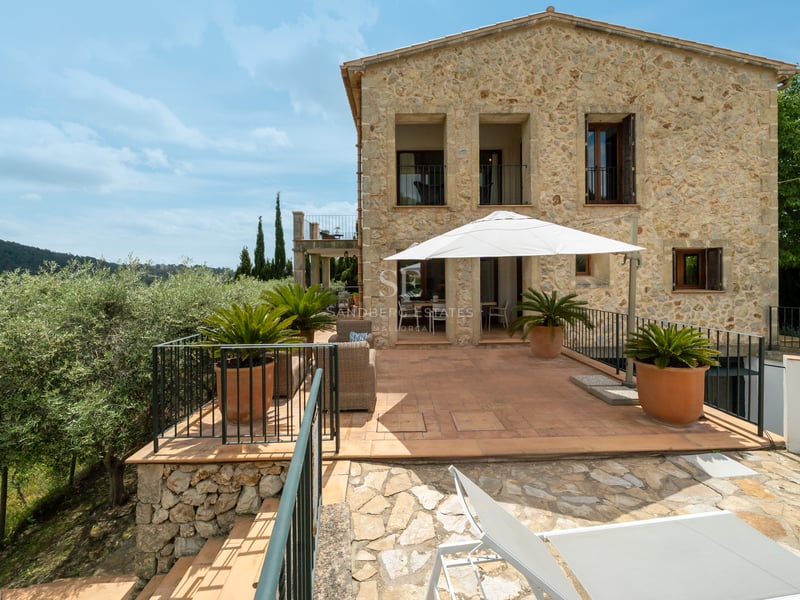 Villa en pierre naturelle avec grande terrasse en terre cuite, parasol blanc et salon de jardin sous un ciel bleu.