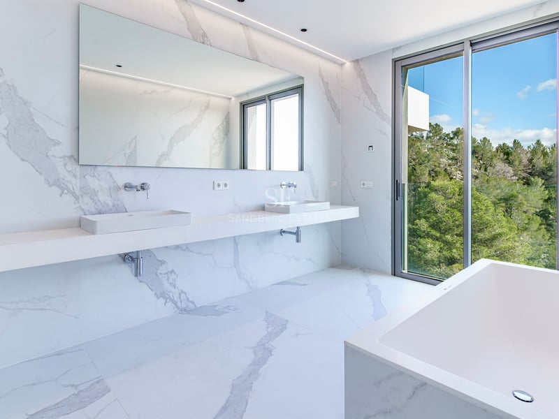 Salle de bain principale lumineuse avec murs et sols en marbre blanc, double vasque, baignoire et vue sur la forêt.