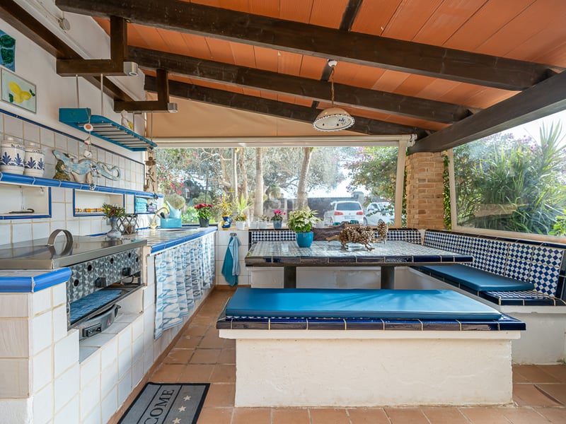 Terrasse couverte avec cuisine d'été en carreaux bleus et blancs, bancs intégrés et plafond à poutres en bois.
