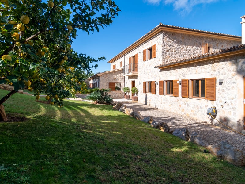 Exteriör av en naturstensvilla med träslutare, grön gräsmatta och citronträd under en klarblå himmel.