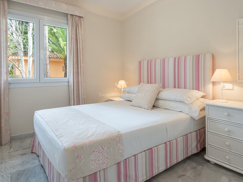 A bright bedroom featuring a double bed with pink textiles, white marble floors, and a window with palm tree views.