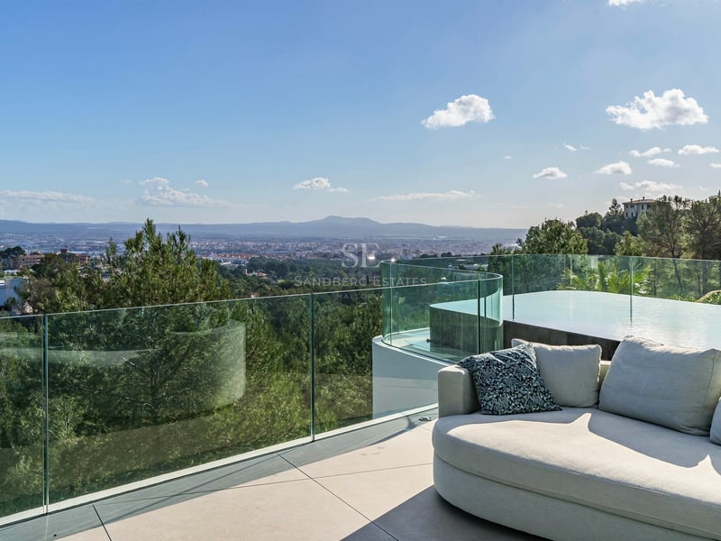 Terrasse moderne avec canapé design, garde-corps en verre et piscine surplombant la ville et les montagnes.