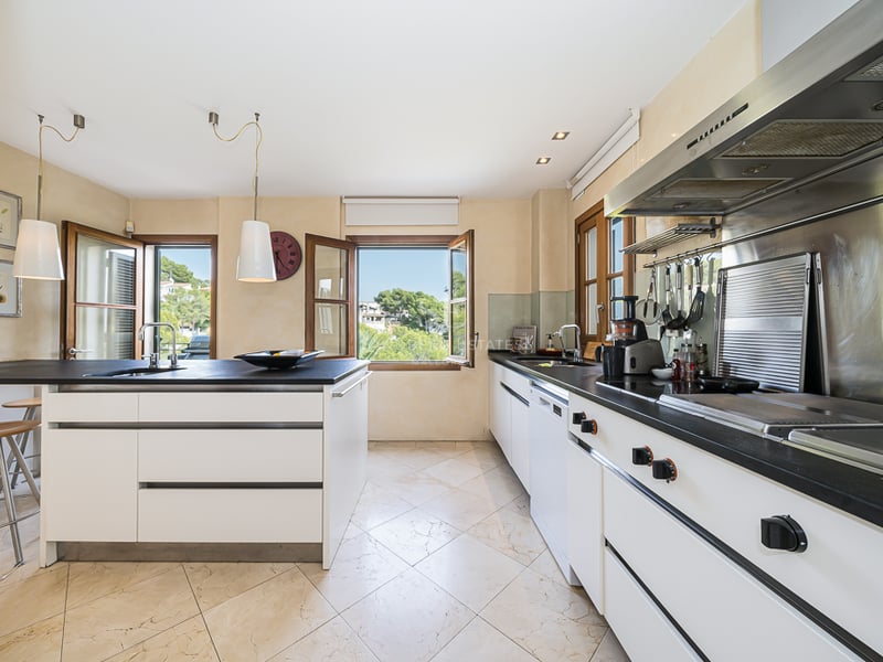 Modernt vitt kök med mörka bänkskivor, klinkergolv och fönster med utsikt över grönska.