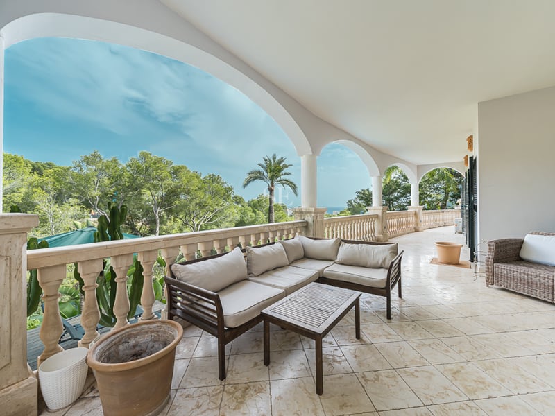 Rymlig terrass med valv, utemöbler, stenbalustrader och utsikt över prunkande grönska och havet.