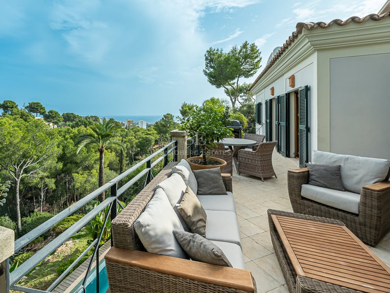 Rymlig terrass med loungemöbler i rotting och stengolv med utsikt över en grönskande dal och havet.