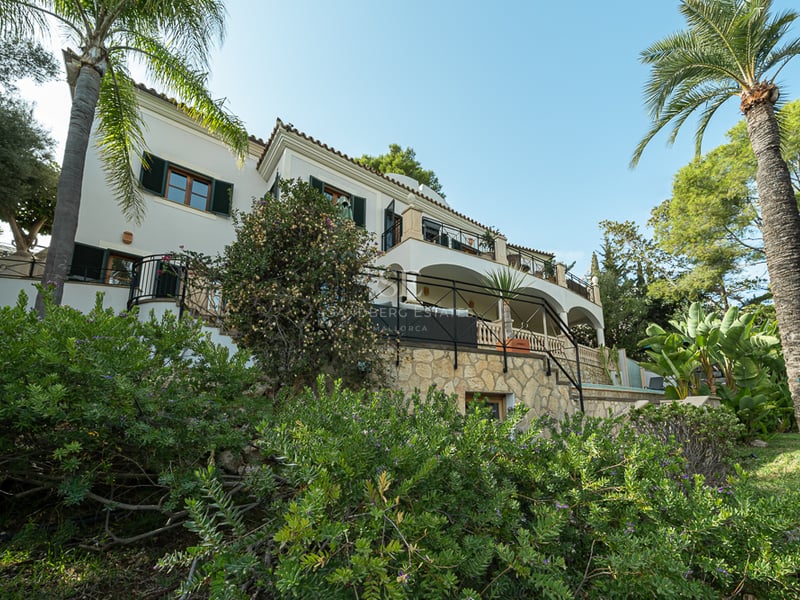 Vit trevåningsvilla med valvterrasser, stenmurar och palmer under en klarblå himmel.