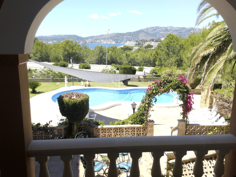 Vista desde un arco de una piscina turquesa, jardín frondoso y el mar Mediterráneo al fondo.