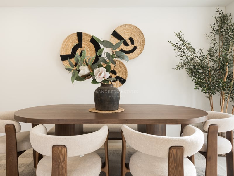 Salle à manger avec table ovale en bois, chaises crème, art mural tissé et un olivier d'intérieur.