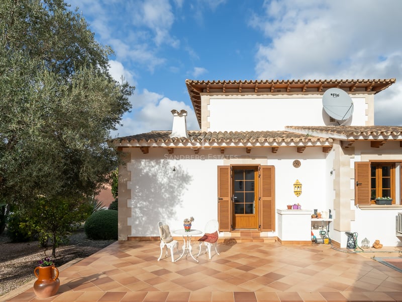 Villa terrace with terracotta tiles, wooden shutters, white outdoor seating, and a large olive tree under a blue sky.