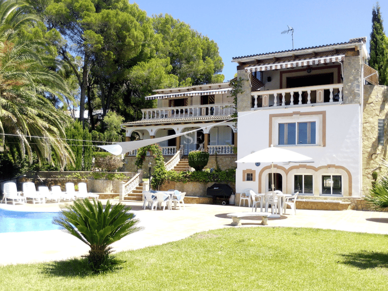 Fachada posterior de una villa blanca de varios niveles con detalles en piedra, piscina, palmeras y terrazas.