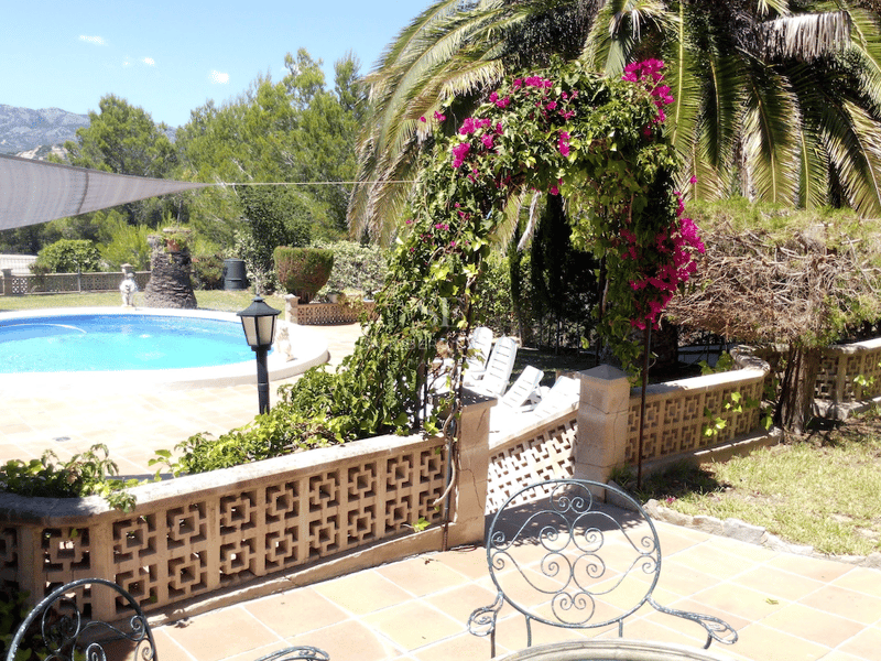 Terraza soleada con piscina circular, baldosas de terracota, un arco de buganvillas y montañas al fondo.