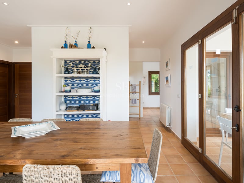 Table à manger en bois massif et chaises en osier dans un intérieur lumineux avec sol en terre cuite et murs blancs.