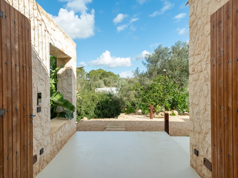 Entré med ljusa naturstensväggar som leder till en lummig medelhavsträdgård under en blå himmel.