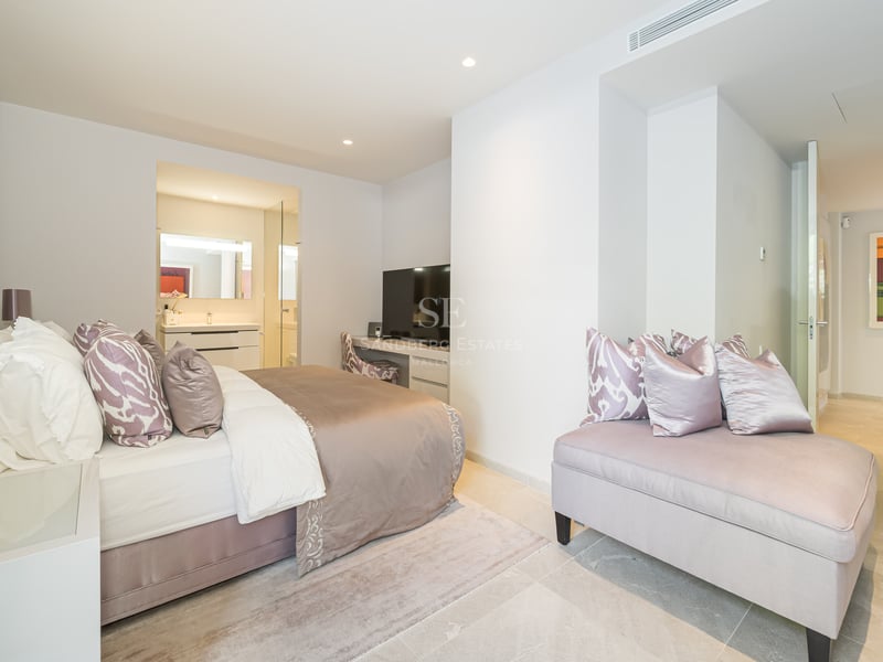 Modernes Schlafzimmer mit Doppelbett, mauvefarbener Recamiere, Schreibtisch und Blick ins Badezimmer.