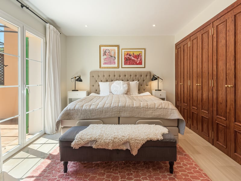 Sun-drenched master bedroom with tufted headboard, large wooden wardrobes, and glass doors opening to a terrace.
