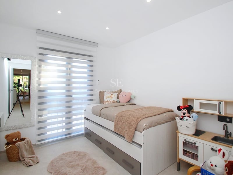 Vitt sovrum med säng med förvaring, helkroppsspegel, zebrapersienner och leksakskök.