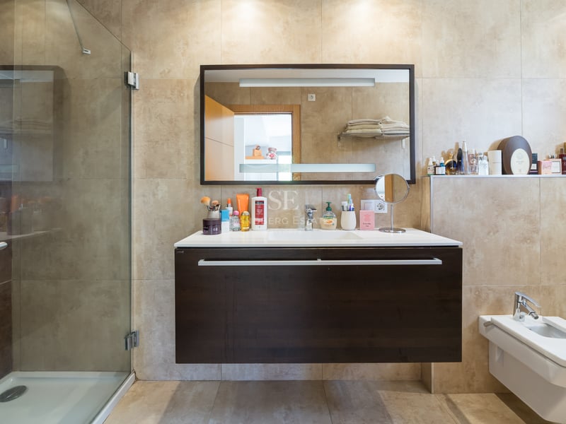 Modernt badrum med glasdusch, mörk svävande kommod och beige kakel.