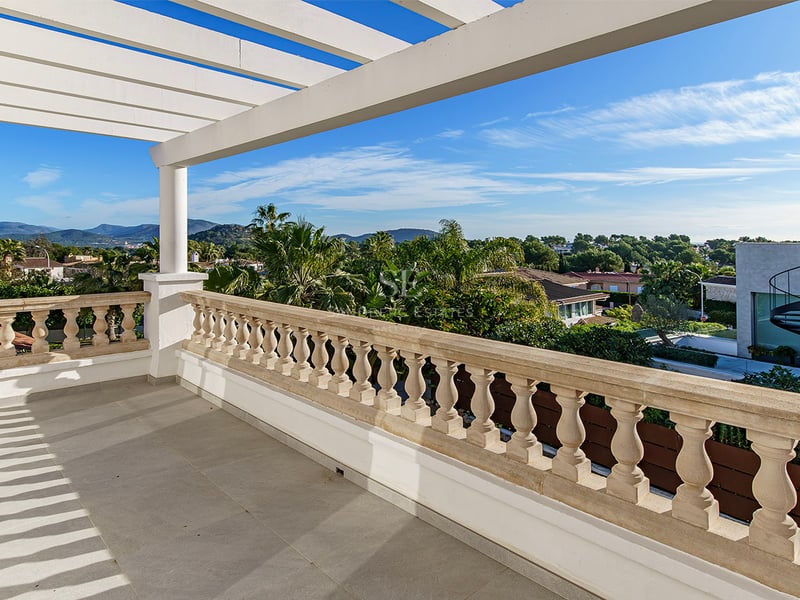 Upphöjd terrass med vit pergola, klassisk stenbalustrad och panoramautsikt över grönska och berg.