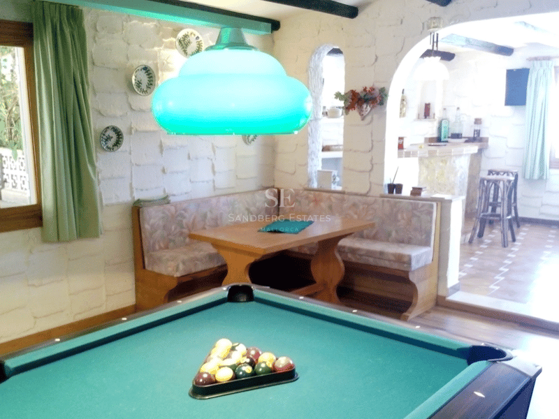 Sala de juegos rústica con mesa de billar, rincón de asientos de madera y paredes de piedra blanca.