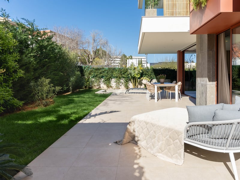 Modern terrace with stone tiling, outdoor dining set, designer lounge chair, and a manicured green lawn under a blue sky.