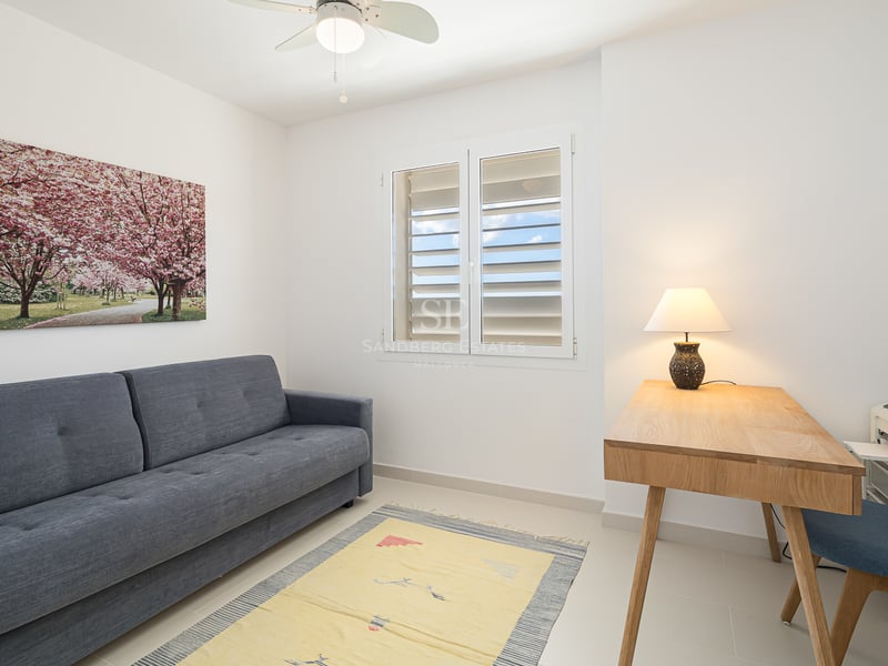 A minimalist room with a grey sofa, wooden desk with a lamp, a blue chair, and a window with white shutters.
