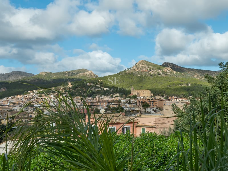 A scenic view of a Mediterranean town with terracotta roofs nestled against lush green mountains under a cloudy blue sky.