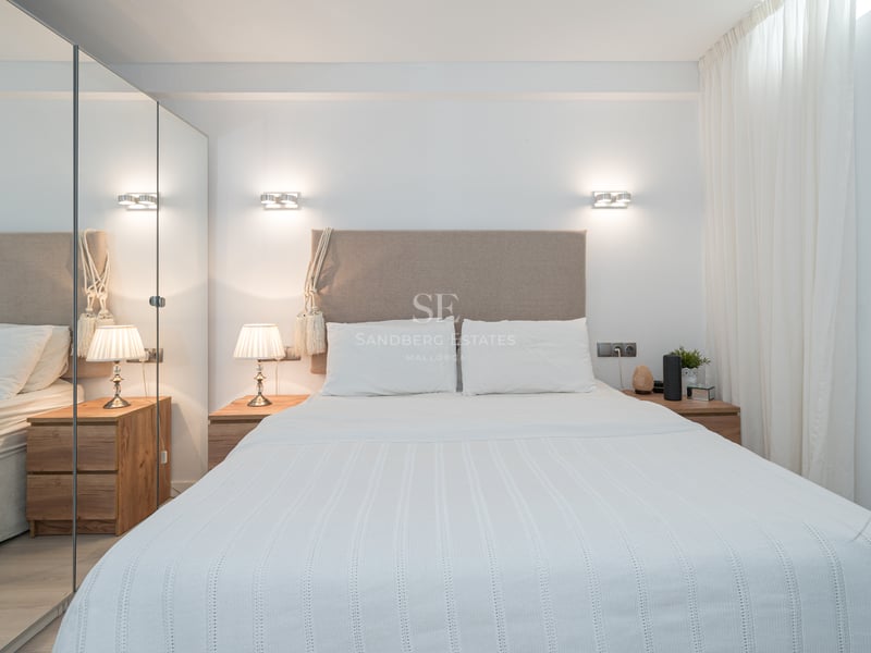 A bright modern bedroom featuring a beige upholstered headboard, white linens, and large floor-to-ceiling mirrored closets.