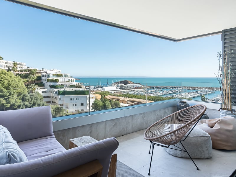 Überdachte Terrasse mit lavendelfarbenem Sofa, Korbstuhl und Glasgeländer mit Blick auf den Yachthafen und das Meer.