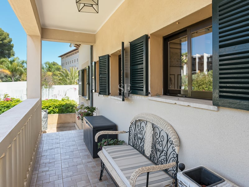 Täckt balkong med smidesbänk, gröna fönsterluckor och utsikt över en grönskande trädgård.