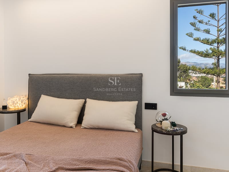 Säng med grå gavel, terrakottafärgat överkast och marmorbord framför ett fönster med utsikt över berg och träd.