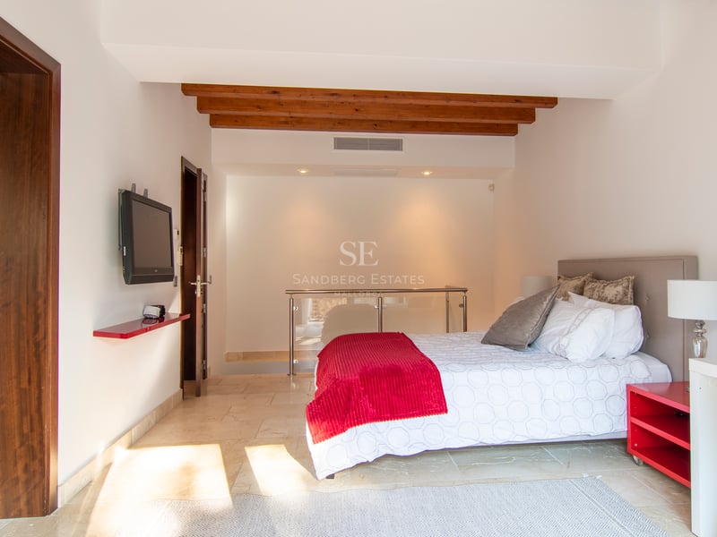 Modernt sovrum med dubbelsäng, röda detaljer, synliga träbjälkar och ljust stengolv.