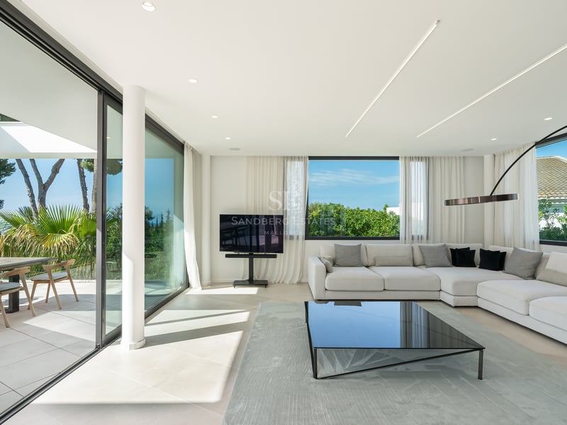 Salon moderne et lumineux avec de grandes baies vitrées ouvrant sur une terrasse avec vue mer et un canapé d'angle blanc.