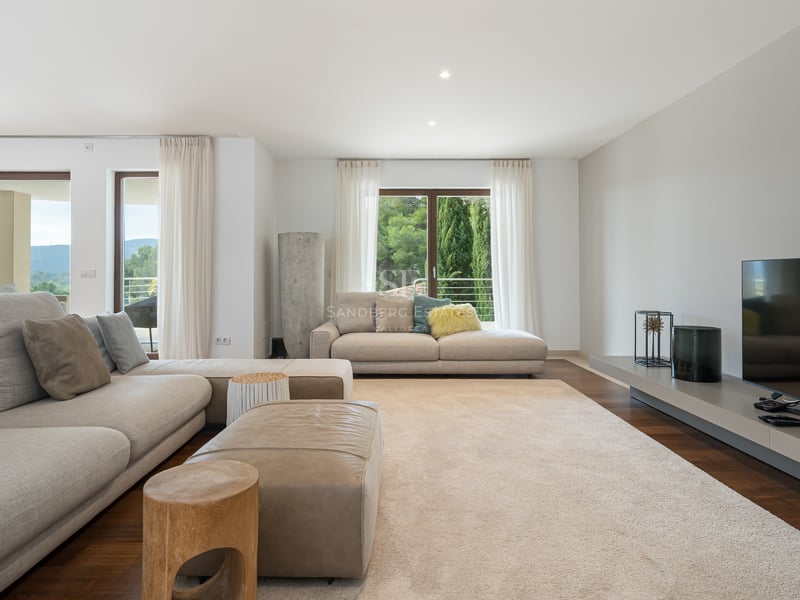 Salon spacieux avec canapés beiges, pouf en cuir, parquet sombre et grandes fenêtres donnant sur des collines verdoyantes.