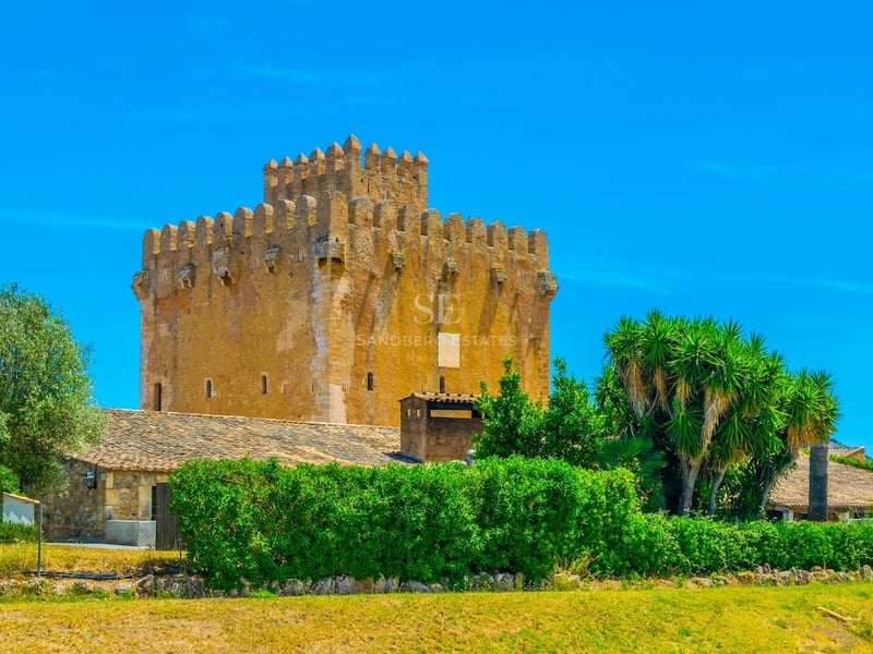 A grand medieval stone tower with battlements under a clear blue sky, surrounded by green hedges and trees.