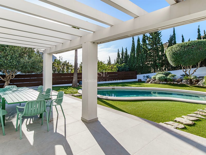 Skuggig terrass med grön matgrupp som vetter mot en pool och anlagd trädgård med cypresser.