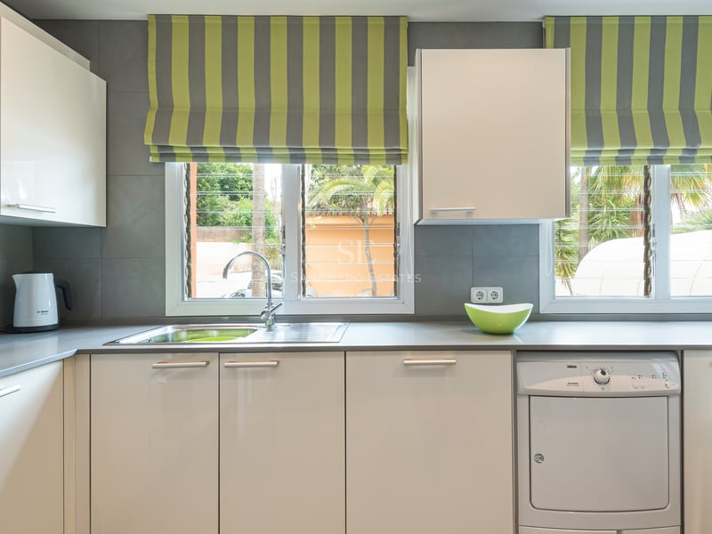 Contemporary kitchen featuring white glossy cabinets, grey countertops, and windows overlooking tropical palm trees.
