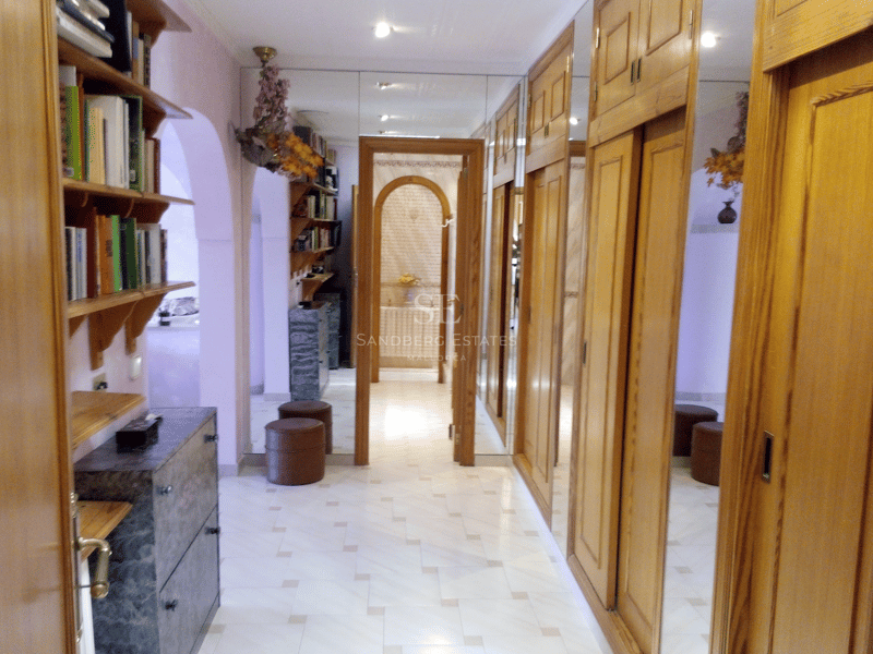 Pasillo con armarios de madera clara, puertas de espejo y suelo de mármol que conduce a un baño.