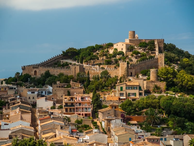 Mediterranean hillside village crowned by a medieval fortress with stone walls under a clear blue sky.