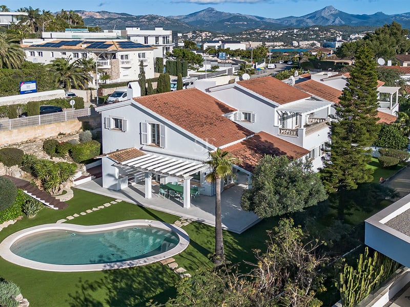 Flygfoto över en vit villa med tegeltak, njurformad pool och grön trädgård mot en bakgrund av berg.