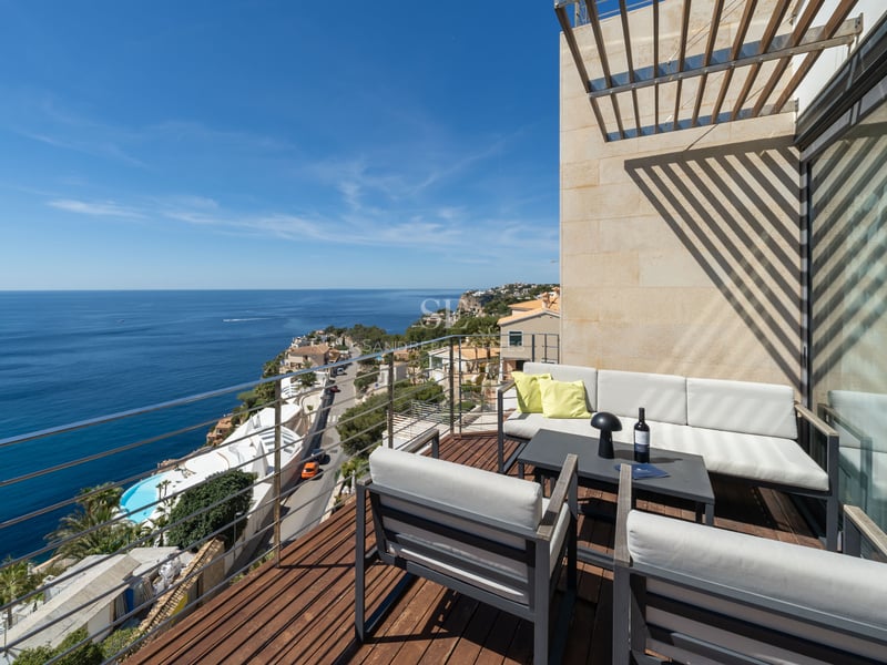 Wooden deck terrace with grey lounge furniture and glass railing overlooking the blue Mediterranean sea under a clear sky.