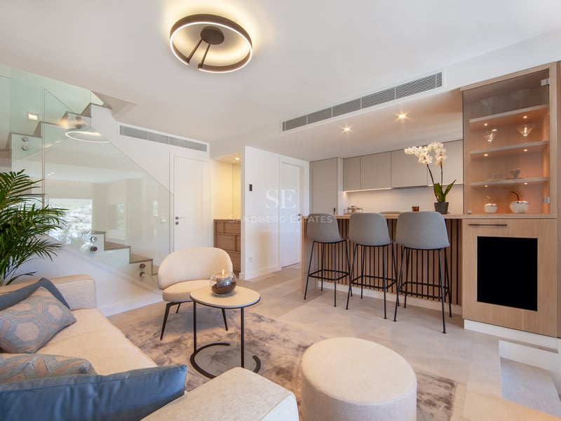 Salon moderne avec canapé crème, escalier en verre et bar à petit-déjeuner en bois avec tabourets gris.