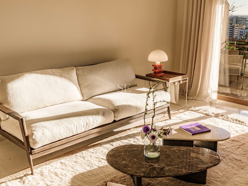 Cremefarbenes Sofa und Marmortische in einem sonnigen Wohnzimmer mit Balkon und Meerblick.