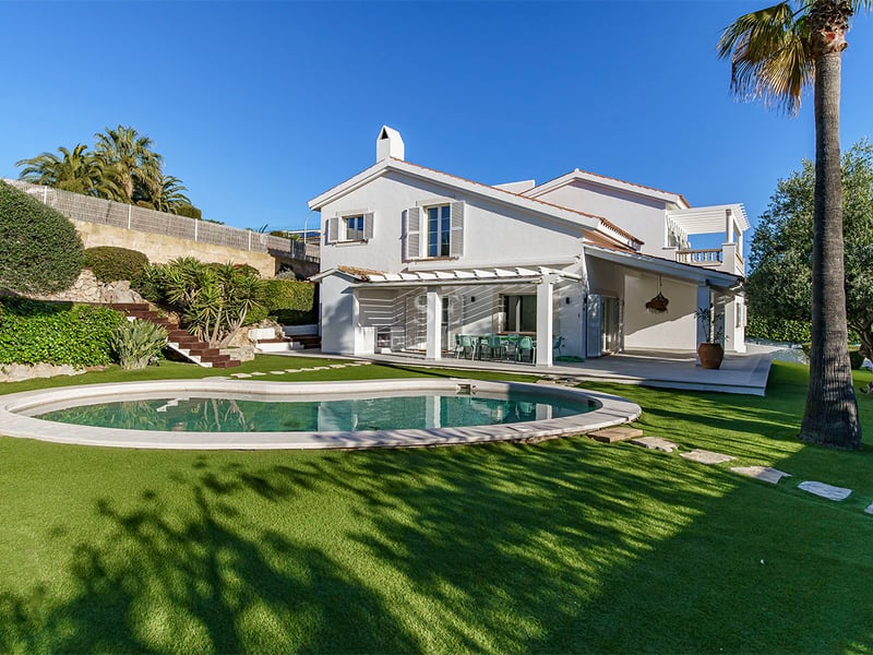 Vit tvåvåningsvilla med njurformad pool, grön gräsmatta och palmer under en klarblå himmel.