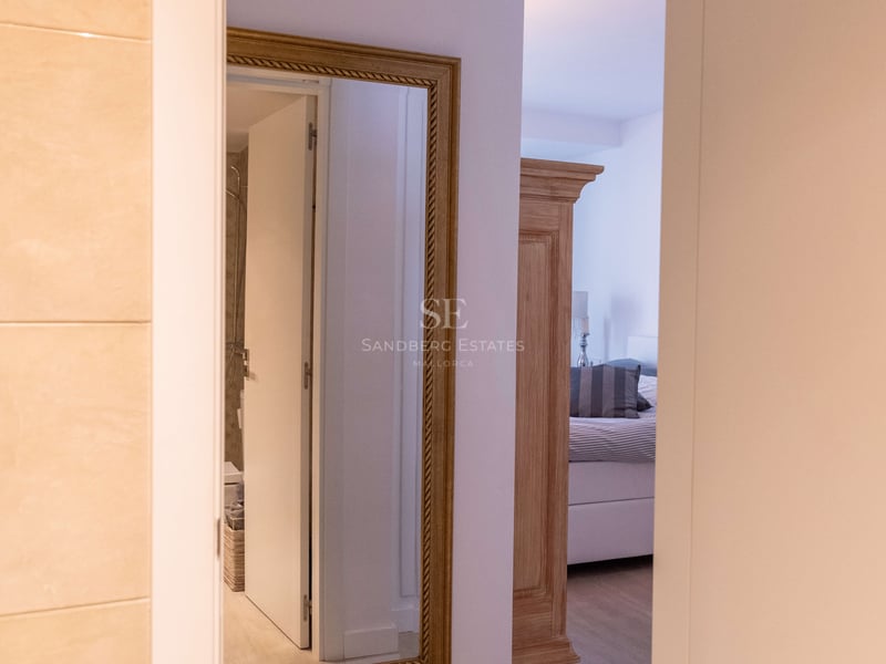 Blick durch eine offene Tür auf einen Flur mit goldgerahmtem Spiegel und Schlafzimmer mit Holzschrank.