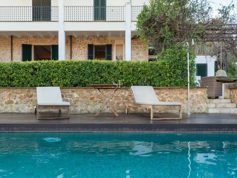 Turkos pool med solstolar i trä framför en stenvilla med gröna fönsterluckor och häckar.