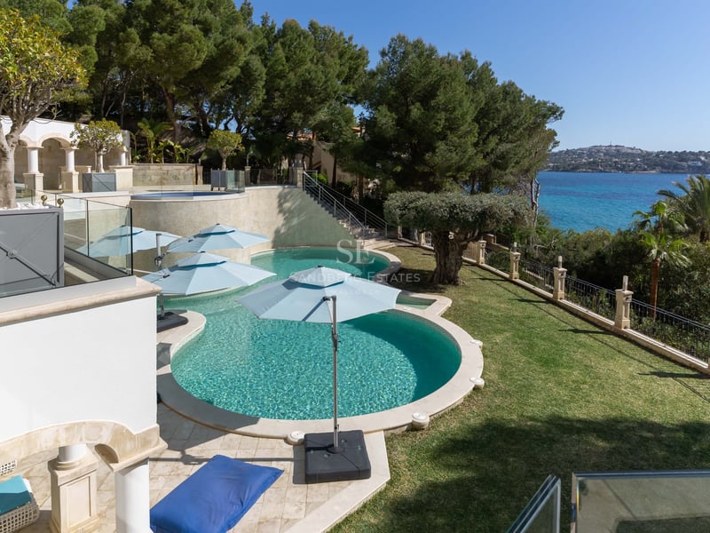 Hochwinkelaufnahme eines geschwungenen türkisfarbenen Pools umgeben von Pinien und Rasen mit Meerblick.